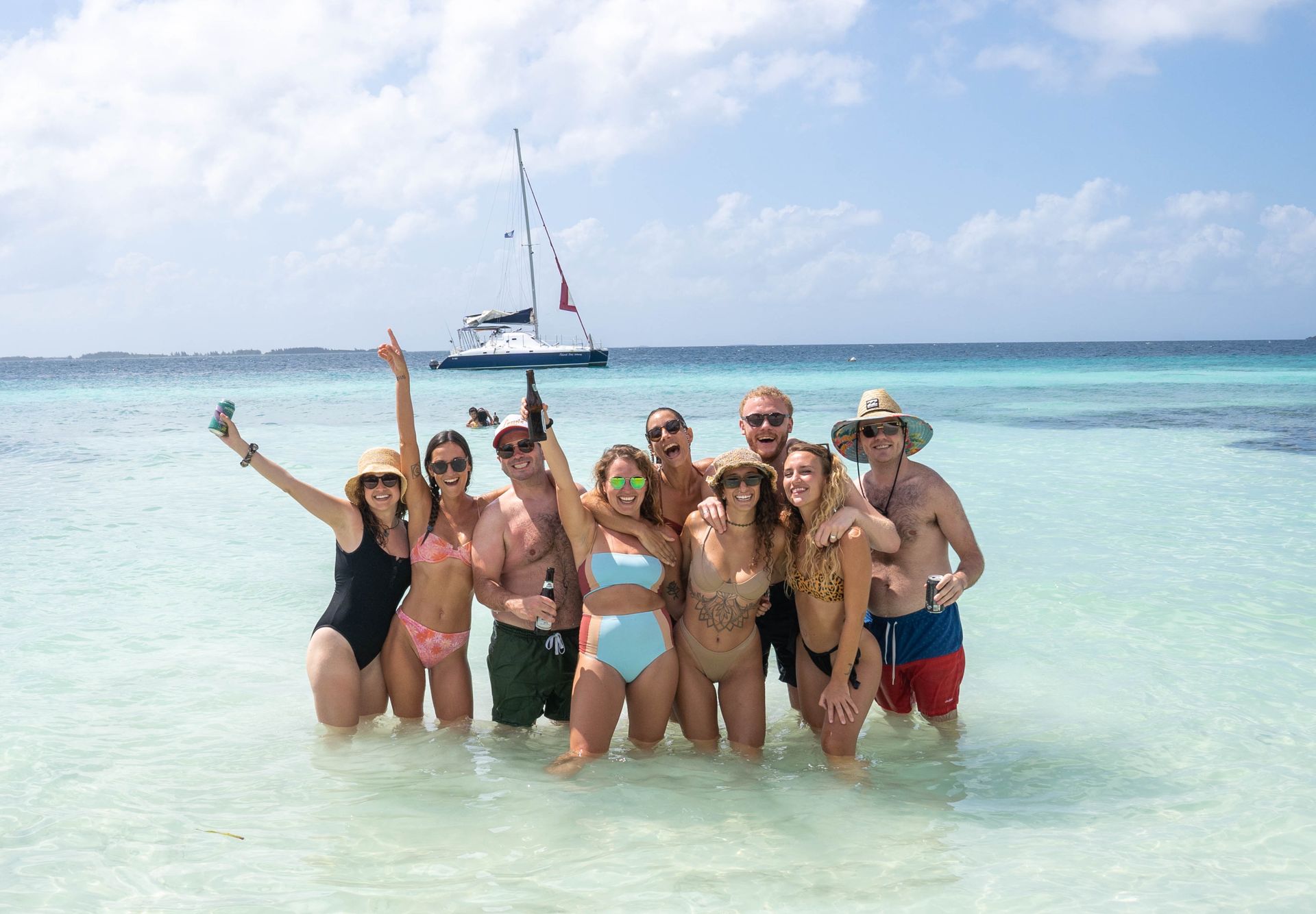 Friends standing together in shallow turquoise water with a sailboat behind them